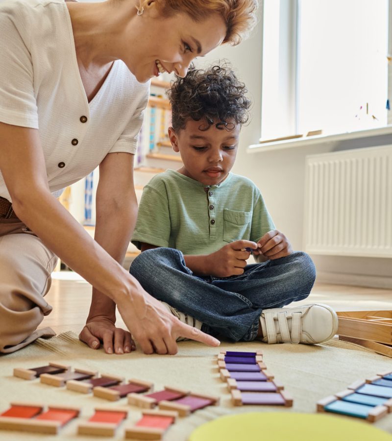 Montessori school, happy teacher pointing at wooden colorful blocks near african american boy, smart