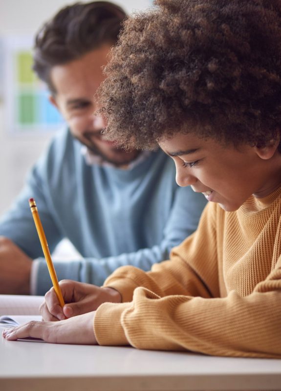 Teacher With Male Student In School Classroom Sitting At Desk Writing In Book Together